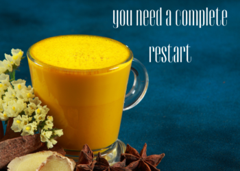 Glass of golden milk surrounded by turmeric, ginger, and spices on a blue background.